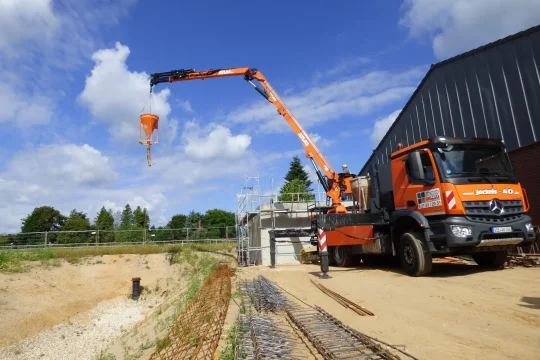 Ein orangefarbenes Mercedes-Benz Kranfahrzeug von Jackels A&O hebt mit einem ATLAS-Kran einen Betontrichter auf einer Baustelle an. Im Hintergrund sind Bauarbeiter und eine Halle zu sehen.