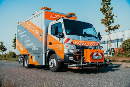 Ein Einsatzfahrzeug von Jackels A&O für die Verkehrsflächenreinigung steht auf einer Straße. Das Fahrzeug ist grau-orange lackiert und mit Reinigungsdüsen an der Front ausgestattet.
